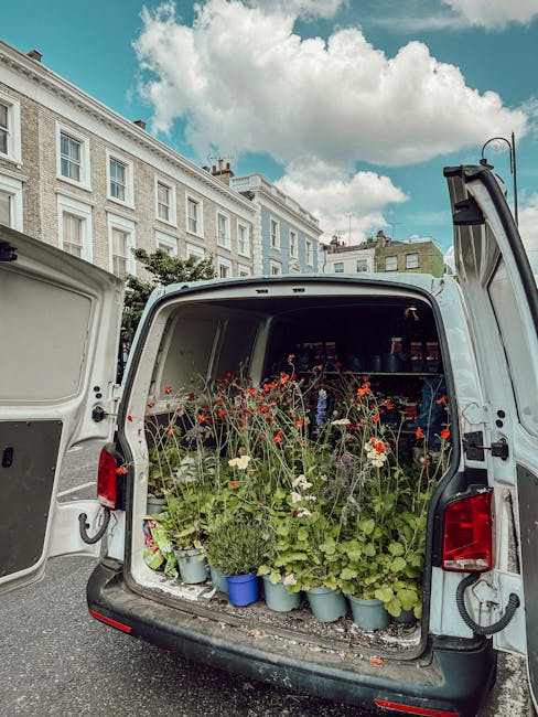 The open rear of a white van filled with potted flowering plants, including red and white blooms, arranged on the vehicle's floor and shelves inside. The plants vary in height and are in small blue an