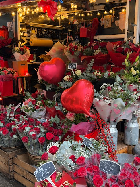 A vibrant display of fresh floral arrangements featuring predominantly red roses, heart-shaped foil balloons, and mixed greenery. The bouquets are wrapped in pink, white, and red tissue paper with som
