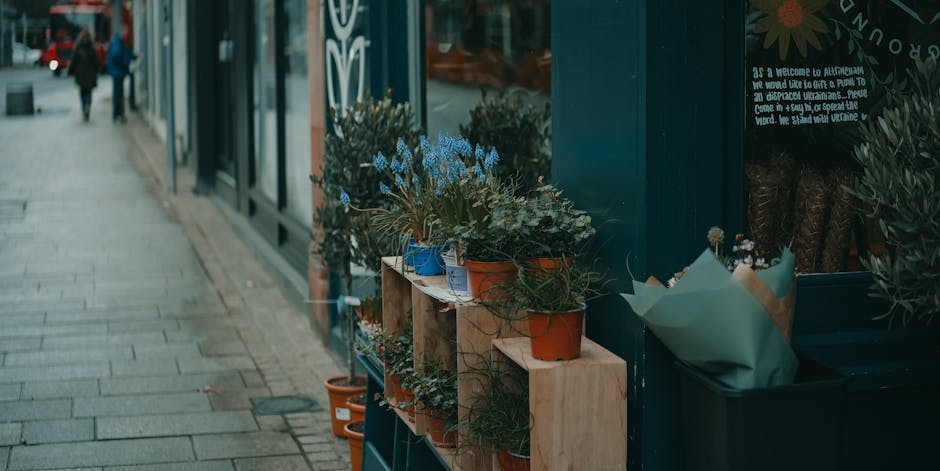 A close-up view of a floral display outside a shop in Cubitt Town, featuring a collection of potted plants and flowers arranged on wooden shelves and in a green container. The arrangement includes pur
