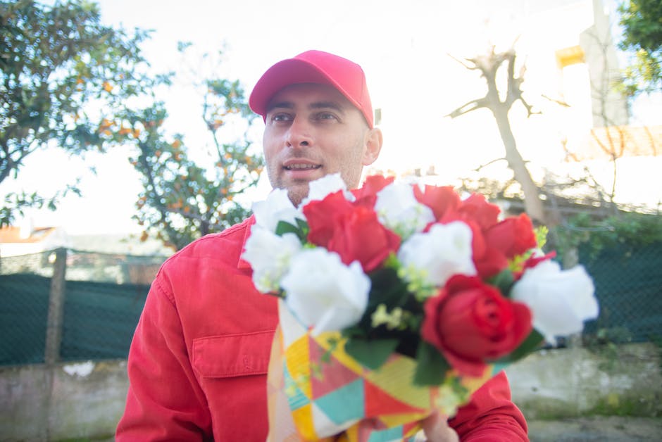 A male flower delivery courier dressed in a red jacket and matching red cap holding a vibrant floral bouquet featuring red and white roses, with visible green foliage, wrapped in colorful paper. The c