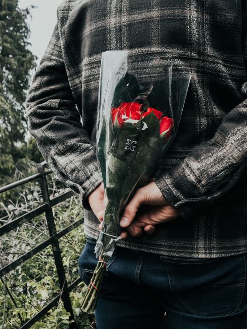 A person wearing a dark plaid jacket is holding a bouquet of fresh red roses wrapped in clear cellophane, with green leaves visible amongst the flowers. The bouquet is arranged with the stems secured 