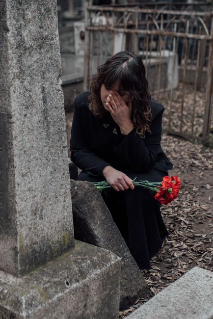 A woman dressed in dark clothing, sitting at a gravesite, holding a bouquet of red chrysanthemums in her right hand while covering her face with her left hand, in a somber outdoor setting with weather
