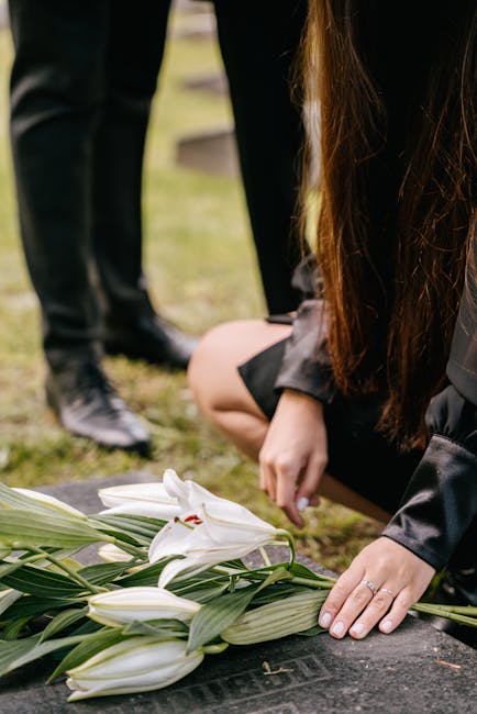 A person wearing a black outfit is kneeling at a outdoor memorial site, gently placing a bouquet of white lilies with long green leaves on a black gravestone. The lilies have large, pristine petals an